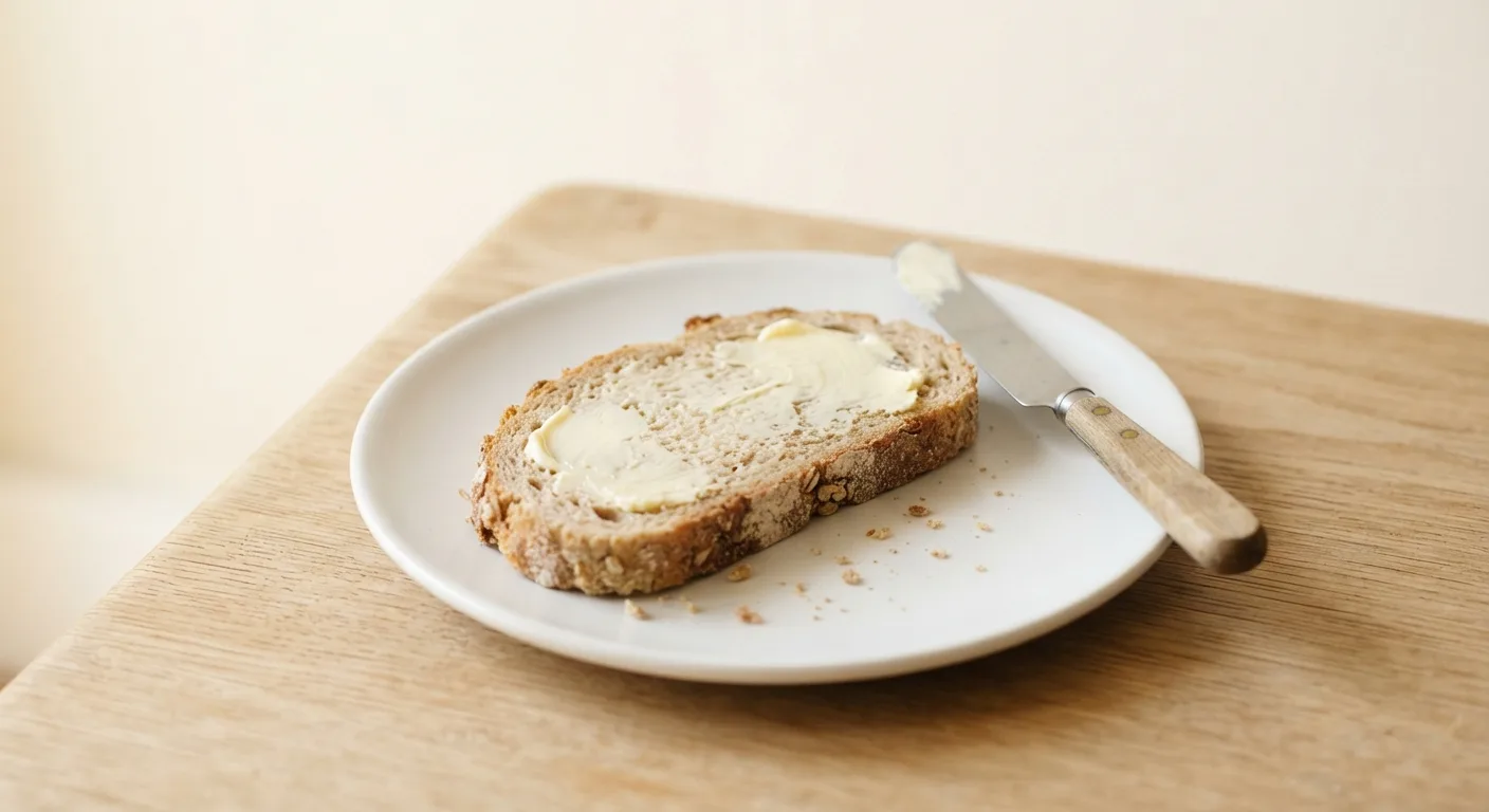 Tranche de pain complet garnie d'une fine couche de beurre, posée sur une assiette au petit-déjeuner